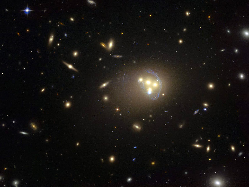 Красивият галактически куп Abell 3827. Странните сини структури, заобикалящи галактиките в центъра на снимките подсказват, че зад тях има и много по-далечни галактики. В момента задълбочено бива изследвано движението на четирите галактики в средата на снимката. По последни данни тъмната материя около една от галактиките не се движи със самата галактика, което значи, че  тя си взаимодейства с неизвестен за учените обект